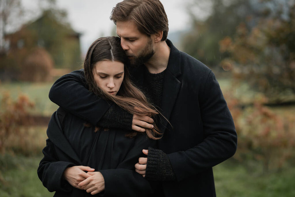 Una joven pareja enamorada de abrigos negros camina en el campo bajo la lluvia. Humor sombrío de otoño. imagen cinematográfica - Foto, imagen