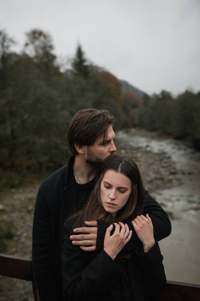 Młoda para zakochana w czarnych płaszczach spaceruje po wsi w deszczu. Jesienny ponury nastrój. obraz kinowy - Zdjęcie, obraz