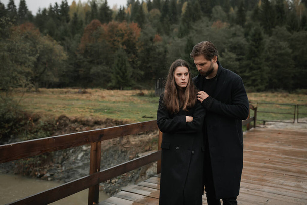 Una joven pareja enamorada de abrigos negros camina en el campo bajo la lluvia. Humor sombrío de otoño. imagen cinematográfica - Foto, imagen