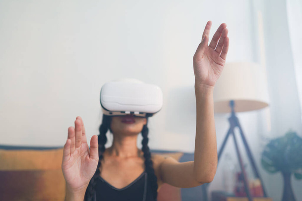Mladá Asiatka s brýlemi pro VR a gesty ve vzduchu, osamělá dívka hrající doma na metaverzní brýle. Selektivní zaměření. - Fotografie, Obrázek
