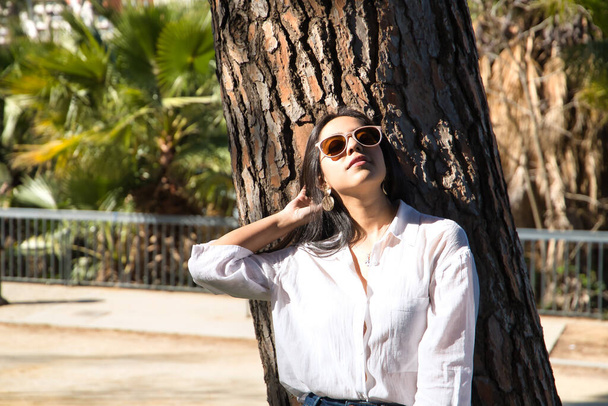 Young and beautiful woman, South American, with sunglasses and white shirt, leaning on the trunk of a tree taking the sun's rays while touching her hair. Concept travel, beauty, fashion, nature. - Photo, image