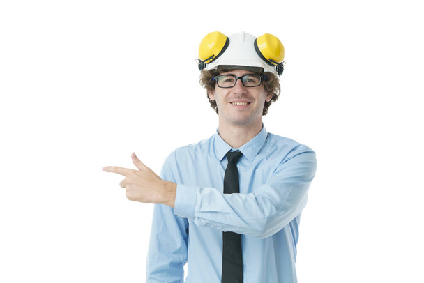 若いエンジニアの男の肖像は、ハード帽子の笑顔を身に着けていると立ちながら、孤立した白い背景の上にフレームの左側に空のスペースに指を指す. - 写真・画像