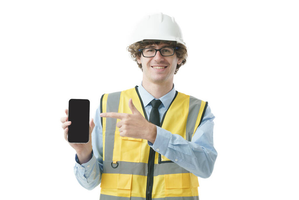 Retrato de capataz ingeniero caucásico con uniforme industrial apuntando dedo a teléfono inteligente con pantalla vacía. - Foto, Imagen