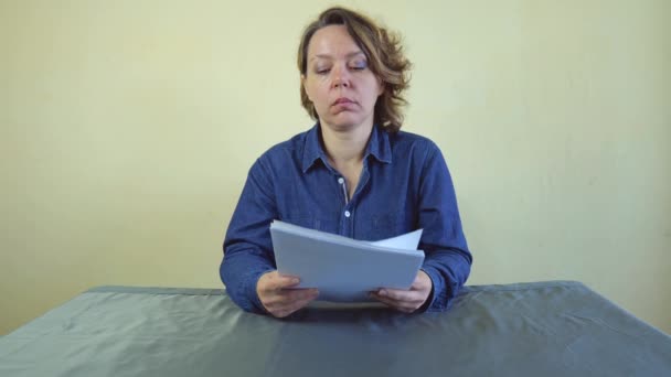 Una mujer de cuarenta años con una camisa de mezclilla sostiene papel blanco en sus manos mientras está sentada en una mesa gris. Retrato en cámara lenta - Metraje, vídeo