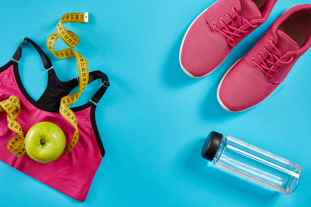 シアンブルーの背景に測定テープを持つスニーカー。黄色、ピンクのスニーカー、女性のスポーツトップと水のボトルでセンチメートル、コピースペース. - 写真・画像