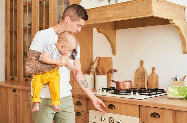 A young Caucasian father and a young son cook together, standing in the kitchen. The father turns on the stove, the oven, holds the child in his arms. The concept of diversity, gender roles. - 写真・画像