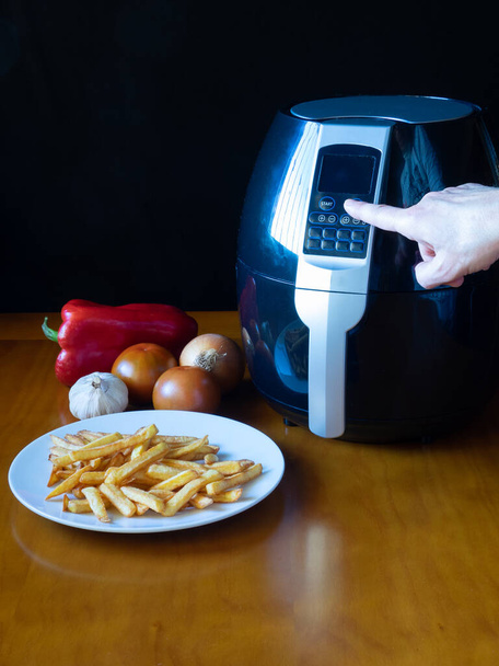 Patates kızartması ve sebzelerle dolu bir masada bir hava kızartma makinesini programlayan bir kadın. Sağlıklı gıda konsepti - Fotoğraf, Görsel