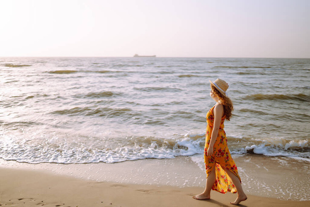 Gyönyörű nő nyári ruhában és kalapban a tengerparton sétál. Utazás, hétvége, pihenés és életmód koncepció. - Fotó, kép