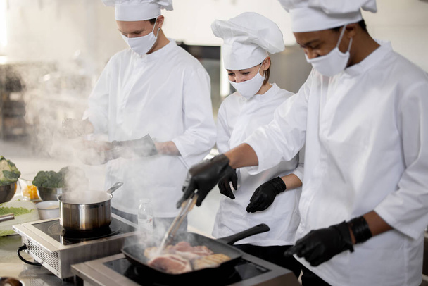 Ein multiethnisches Team von Köchen in Uniform und Gesichtsmasken kocht Mahlzeiten für ein Restaurant in der Küche - Foto, Bild