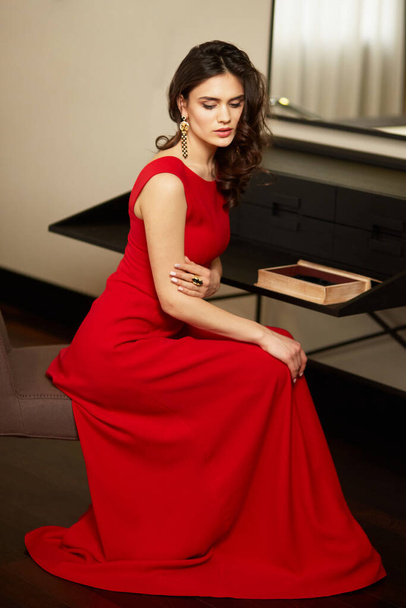 Gorgeous young woman with brown hair in elegant red dress sitting on chair in luxury interior - Foto, Imagem
