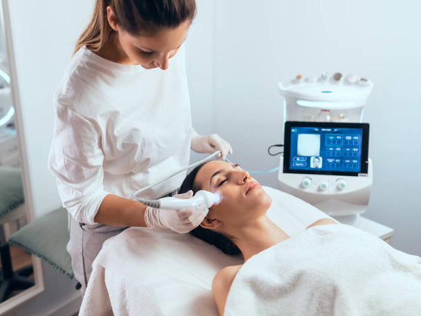 Shot of cosmetologist making ultrasonic cleaning and rejuvenation the face to beautiful woman on the spa center. - 写真・画像