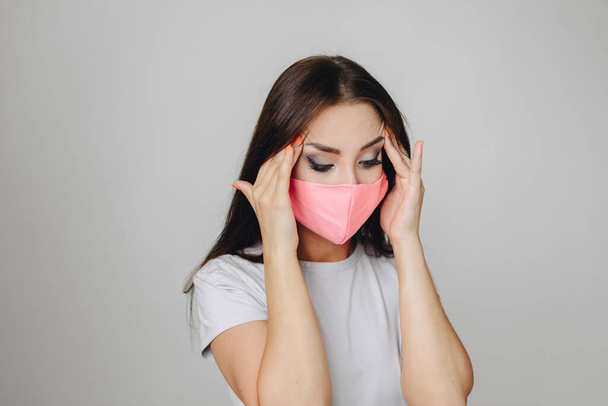 A brunette in a white T-shirt on a white background in a pink protective mask stands holding her hands at her temples looking down. The concept of illness, flu. - 写真・画像