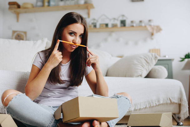 A brunette sits on the floor near the sofa among cardboard boxes, parcels and holds a pencil near her lips while looking at the camera. - Фото, изображение