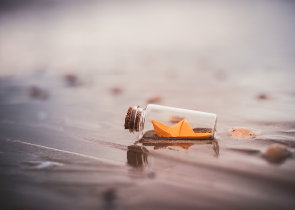 Miniature origami ship in bottle standing on sand at sunrise - Photo, Image