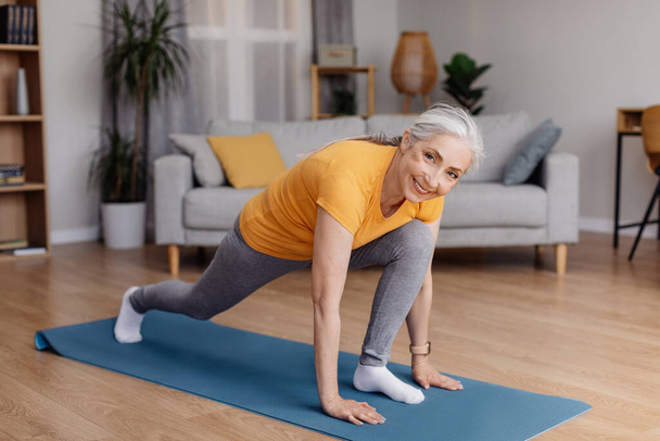 Atletik yaşlı kadın bacaklarını esnetiyor, koşucular yoga pozisyonu alıyor ev antrenmanında, oturma odasında egzersiz yapıyor. - Fotoğraf, Görsel