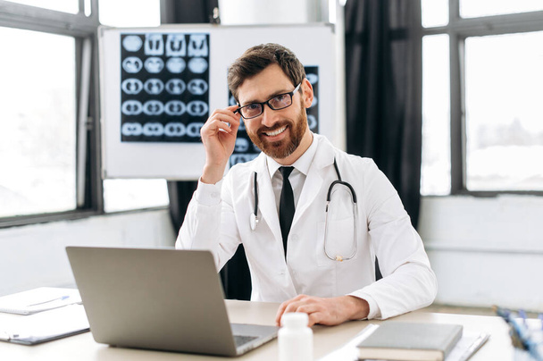 Portret pewnego siebie białego lekarza w płaszczu medycznym, w okularach i stetoskopie, siedzącego w gabinecie lekarskim, patrzącego w kamerę, uśmiechniętego przyjaznego - Zdjęcie, obraz
