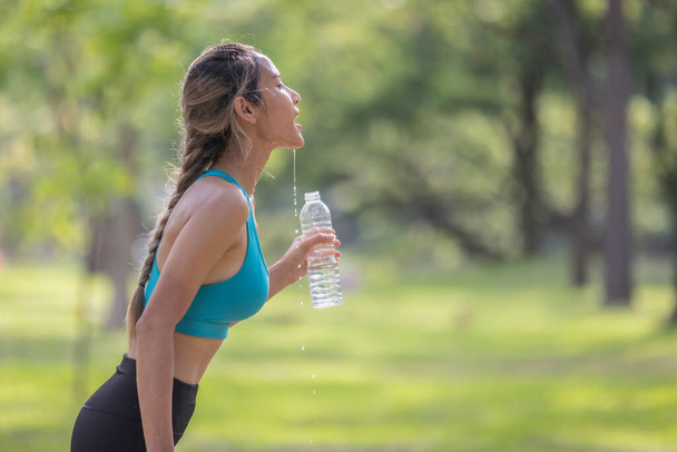 Счастливая молодая женщина улыбается в сторону во время питья воды из бутылки после тренировки опираясь на дерево - Фото, изображение