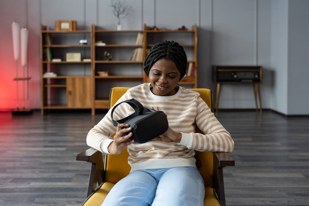 Joven mujer negra emocionada sentada en un sillón en casa, mirando curiosamente los auriculares VR en las manos - Foto, imagen
