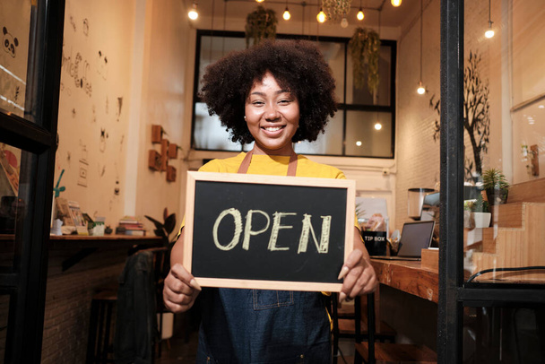 Jedna Afroamerykanka startująca barista stoi przy zwykłych drzwiach kawiarni, spogląda w kamerę i pokazuje otwarty znak, szczęśliwy i wesoły uśmiech z pracą w kawiarni i nowymi przedsiębiorcami. - Zdjęcie, obraz