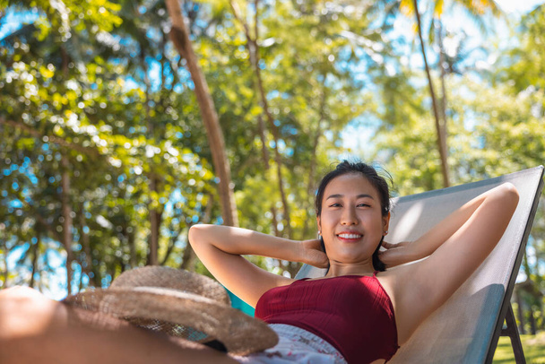 Model asijské ženy na sobě červené plavky a slaměný klobouk ležící na lehátku na slunci u moře. Krásná dívka relaxuje v nekonečném luxusním resortu. Lázně, wellness. - Fotografie, Obrázek