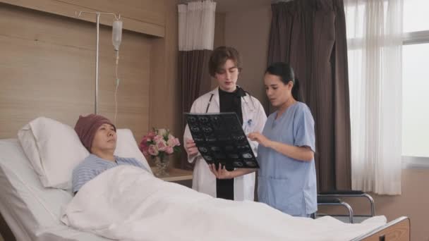 Kaukázusi nő orvos egyenletes diagnózis magyarázza röntgen film ázsiai radiológus és helyreállítási férfi beteg fekvőbeteg szobában egy kórházi kórteremben, orvosi klinika, rák vizsgálat konzílium. - Felvétel, videó