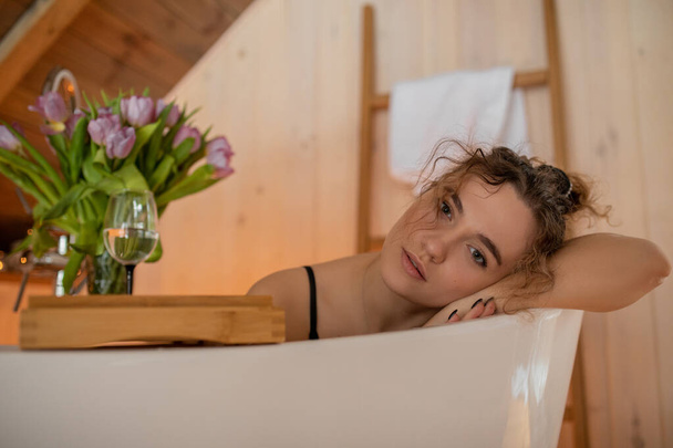 Portrait ofattractive brown-haired woman with curly hair resting, resting her head on her elbow on side of bathroom. Natural wood. Skin and hair care. Luxury and pleasure. Healthy lifestyle. - Photo, Image