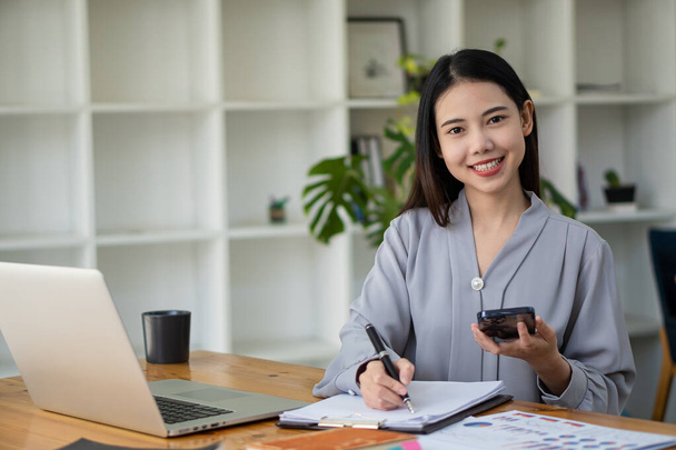 Asian businesswoman holding a smartphone analyzing a report pointing to a graph with a pen, laptop, and calculator. in workstation accountant telemarketing ideas ecommerce free online marketing - Фото, изображение