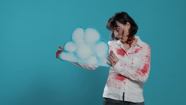 Mujer zombie espeluznante sosteniendo el arte de la nube de cartón mientras está de pie sobre fondo azul. - Metraje, vídeo
