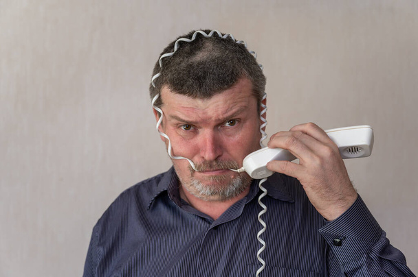 Porträt eines erwachsenen Mannes, der sich ein Telefonkabel um den Kopf wickelt. Ein Mann in einem blau gestreiften Hemd mit einem verdutzten Gesicht. Der Mann beißt auf den weißen Draht. Innenräume. - Foto, Bild