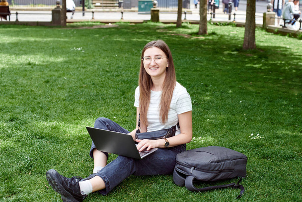 Młoda, ładna kobieta pracuje z laptopem na trawie. Praca w parku na laptopie - Zdjęcie, obraz