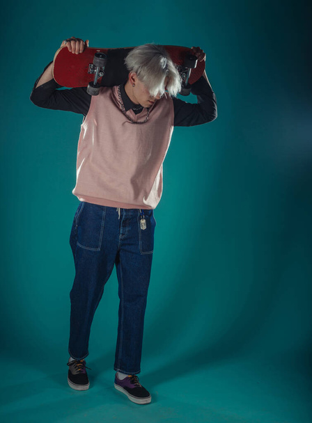 Молодой взрослый мужчина в повседневной одежде смотрит в камеру, держит скейтборд возле лица и стоит в студии на синем фоне - Фото, изображение