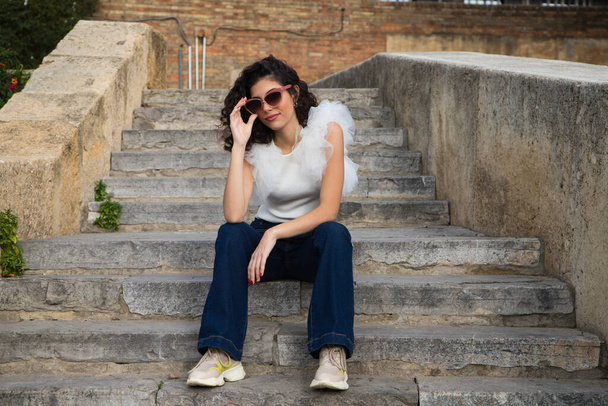 hermosa joven con el pelo oscuro y rizado sentado en una escalera en un parque. La mujer usa gafas de sol y hace diferentes expresiones. Expresiones y concepto de estilo de vida - Foto, imagen