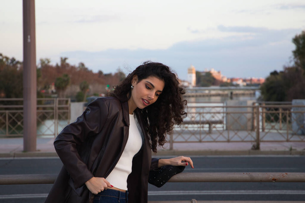 Young and beautiful woman with dark and curly hair is sightseeing in Seville. In the background part of the Mediterranean style European city in the golden hour. Tourism and holidays concept. - Foto, imagen