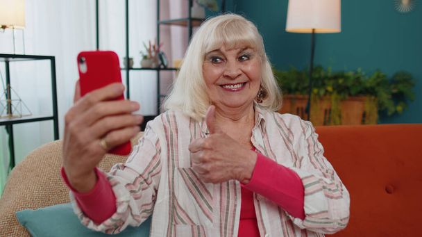 Glückliche Senioren-Großmutter hält Smartphone in der Hand und schaut sich Handy-Videotelefonie im Internet an und macht Selfie nach Hause - Foto, Bild