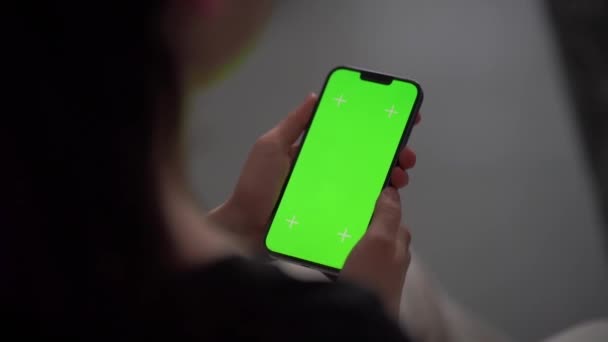 A girl holds her phone while in the living room. Close up of a woman using a smartphone with one hand. Green screen mockup for keying. - Footage, Video