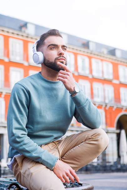 mladý muž sedí na ulici a poslouchá hudbu s bezdrátovými sluchátky - Fotografie, Obrázek