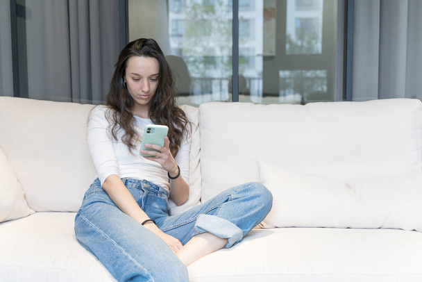 Mujer europea joven que utiliza el teléfono inteligente en el hogar ambiente relajado en el sofá en tiempo libre. Internet, concepto tecnológico. - Foto, Imagen