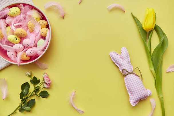 Easter post card concept. Pink bowl with colorful eggs, pink feathers, spring flowers and handmade cute pink bunny. View from above. Copy space. - 写真・画像