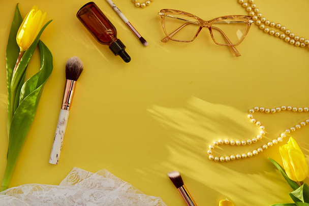 Pastel feminine flat lay with pearls, makeup brushes, glasses, serum dropper and lace. Yellow tulips with trendy shadows. Beauty card concept. Copy space. - Photo, Image