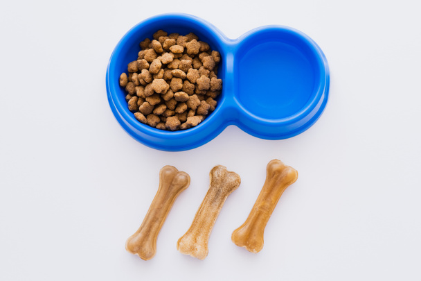 vista superior de las golosinas para mascotas en forma de hueso cerca de un tazón con agua y comida seca para mascotas aislada en blanco - Foto, Imagen