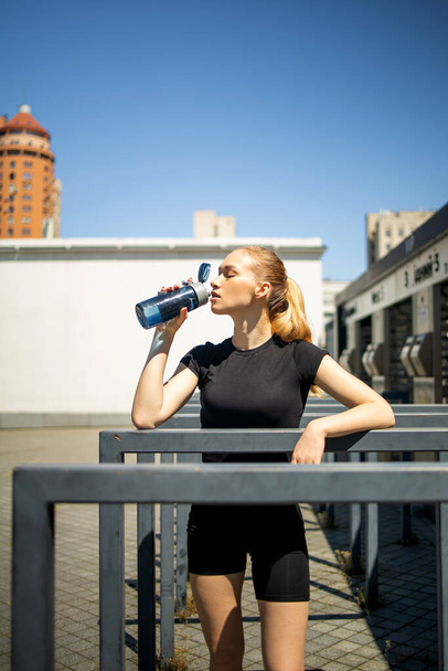Athletic blonde woman in sportswear drinking a protein shake or a bottle of water after a workout outdoors. The concept of sports and recreation. - Foto, Imagem