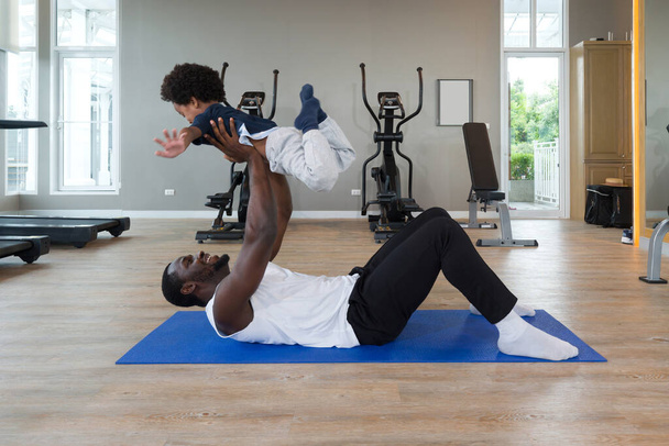 Young short curly black hair father  with moustache and beard in sportswear playing with his son after finish exercise routine on yoga mat. Cardio machines are on the background at the gym. - Photo, Image