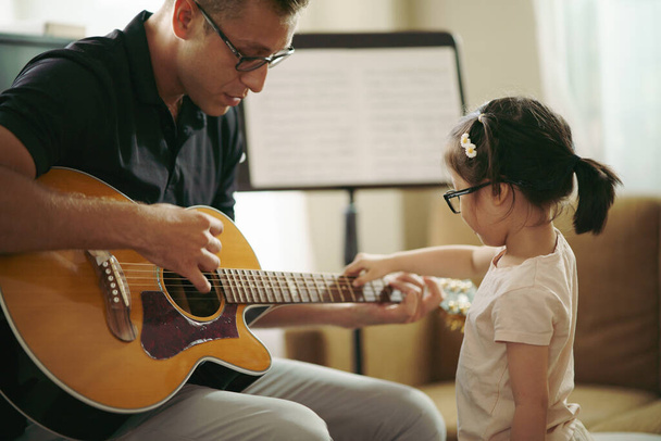 Сцена, где малыш исследует музыкальный инструмент, прелестная маленькая девочка с учителем музыки, играющим на гитаре, пока она экспериментирует со звуком гитарных струн. развитие детского мозга. Возвращение в школу. - Фото, изображение