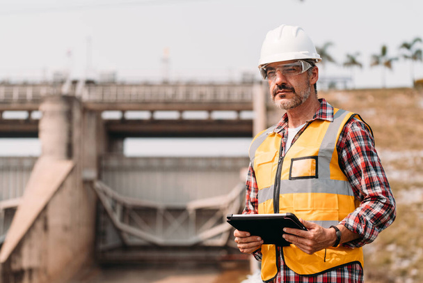 Portrait of power engineer wearing safety jacket and hardhat with tablet working at outdoor field site that have water spillway  of hydro power dam electrical generator at the background. - Photo, Image