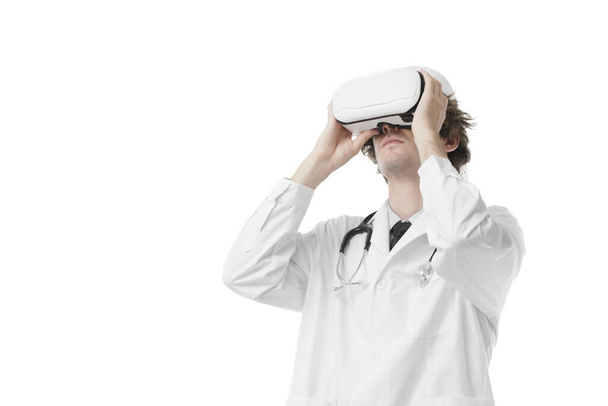 Γιατρός που φοράει ακουστικά εικονικής πραγματικότητας και κοιτάζει πάνω από απομονωμένο λευκό φόντο. Έννοια εικονικής πραγματικότητας ιατρική τεχνολογία υγειονομικής περίθαλψης. - Φωτογραφία, εικόνα