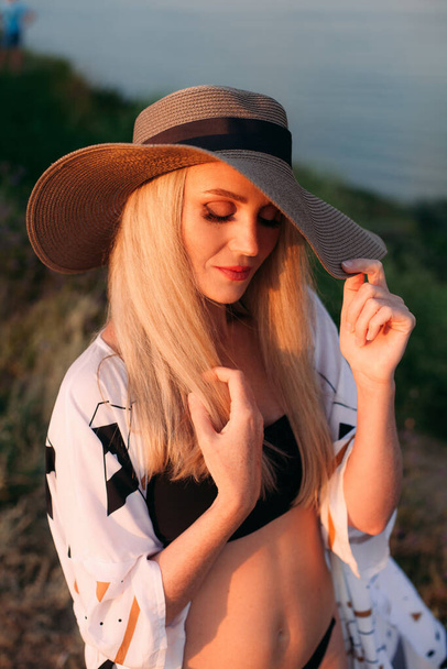 Portrait of a beautiful blonde girl with long hair in a black swimsuit, a light pareo and a hat, near the ocean, at sunset. - Foto, Imagem