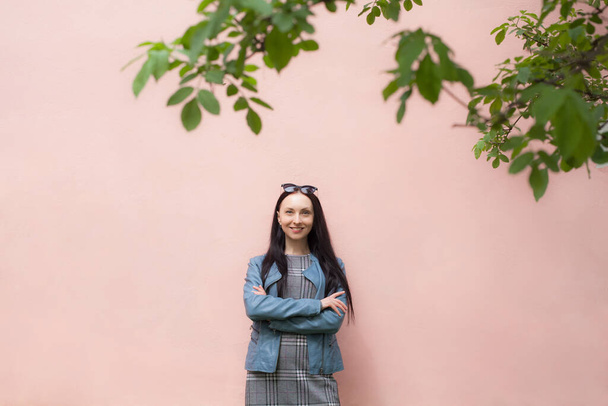 A positive girl with a beautiful smile in a gray dress and a blue denim jacket, with glasses on her head, stands near a pink clean wall, against her background there are several branches from trees. - Foto, afbeelding