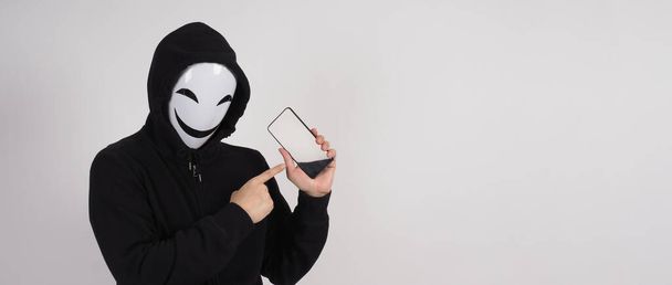 Ανώνυμος χάκερ και μάσκα προσώπου με smartphone στο χέρι. Άντρας με μαύρη κουκούλα που κρατάει και χρησιμοποιεί κινητό τηλέφωνο σε λευκό φόντο. Αντιπροσωπεύουν το κυβερνοέγκλημα δεδομένα hacking ή κλοπή προσωπικών δεδομένων έννοια - Φωτογραφία, εικόνα