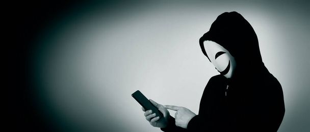 Hackeador anónimo y máscara facial con smartphone en la mano. Camisa hombre de capucha negra sosteniendo y usando teléfono móvil sobre fondo blanco. Representar el cibercrimen piratería de datos o robo de datos personales concepto - Foto, imagen