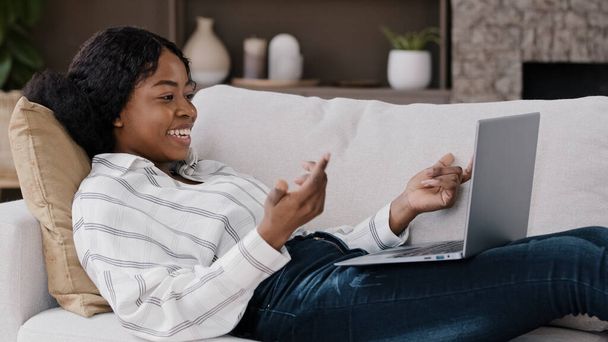 African woman lying on sofa at home look at laptop screen greeting hello make video call talking having remote conversation with friend family living abroad. American female communicates by conference - 写真・画像